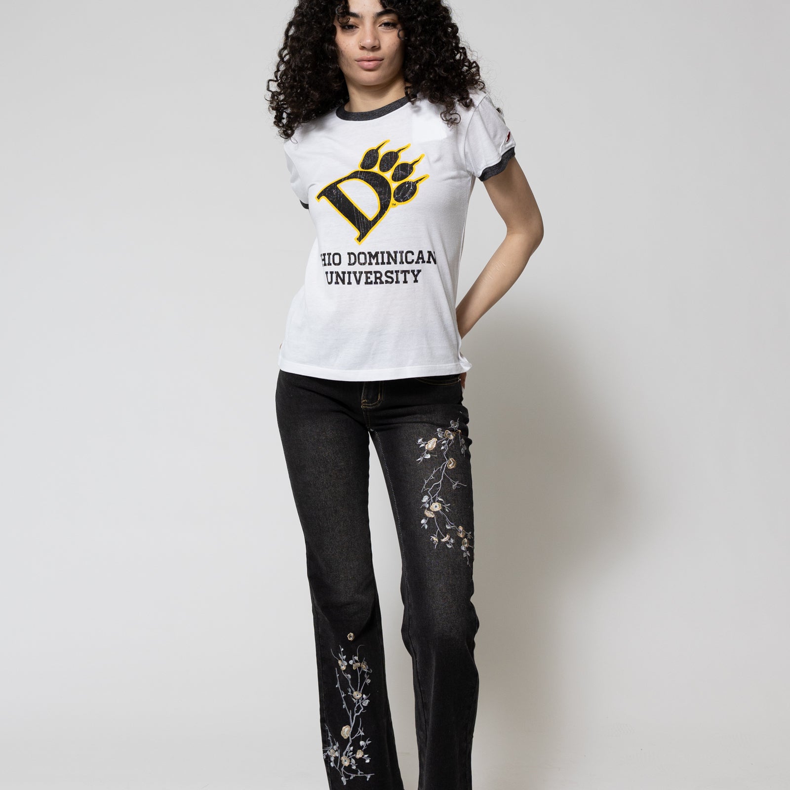Person wearing a white t-shirt with a logo and text, black jeans with floral designs, and black sneakers on a white background.