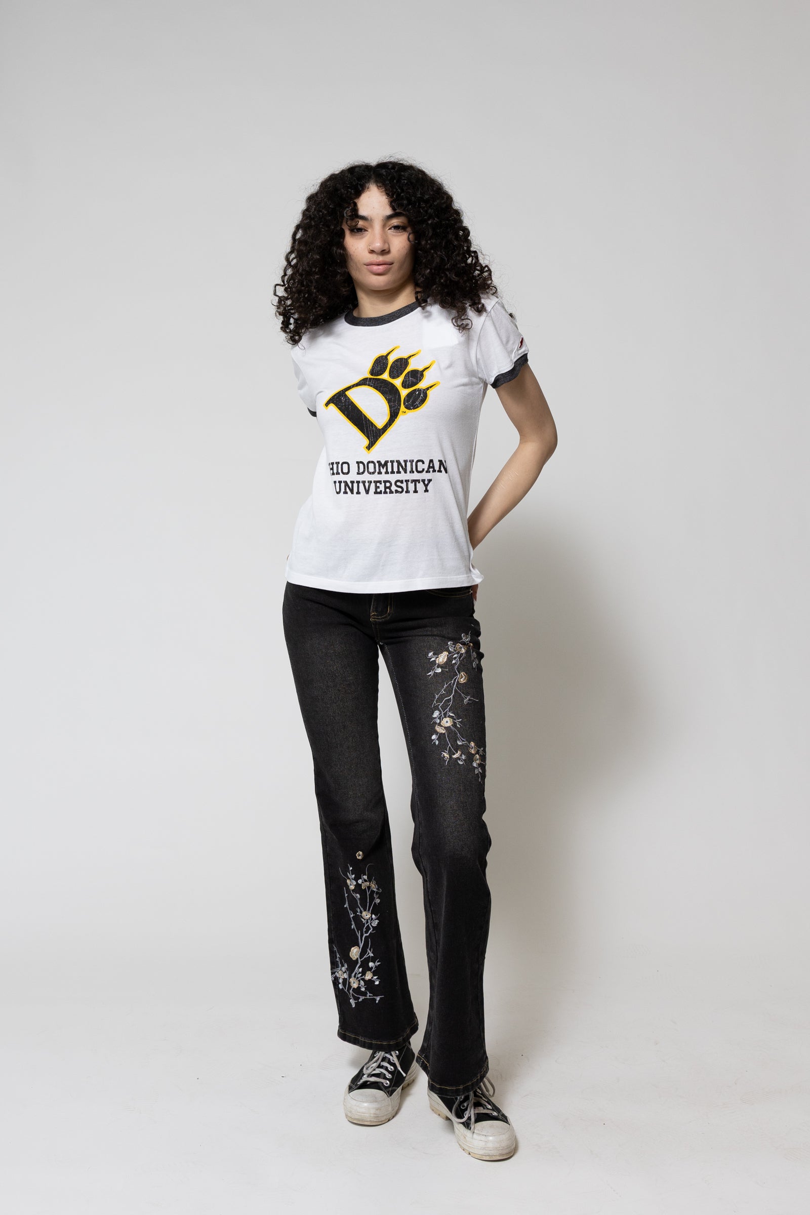Person wearing a white t-shirt with a logo and text, black jeans with floral designs, and black sneakers on a white background.
